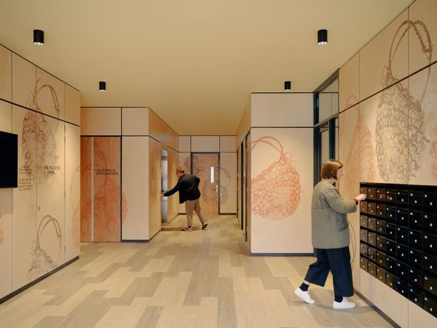A contemporary apartment or commercial building entry lobby with a long corridor, pale timber-look flooring, warm beige ceilings, and large cream wall panels decorated with delicate line-art botanical and abstract graphics. A black multi-compartment mailbox wall occupies the right side, while recessed and surface-mounted black downlights provide contrast. Glass doors and glazed partitions lead further into the space, with people using the entry area.