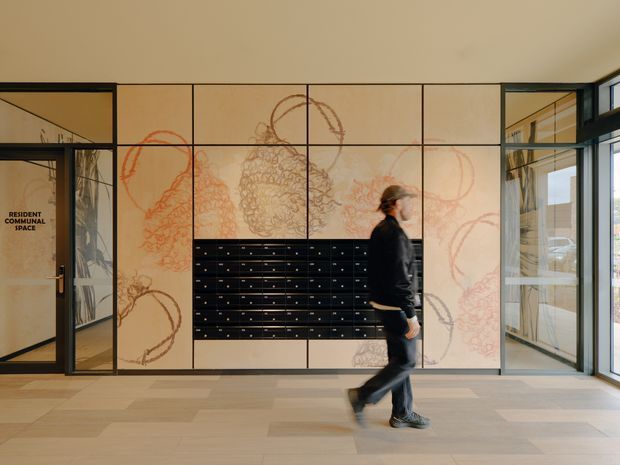 A contemporary communal lobby or entry space with pale timber wall panels featuring large abstract line artwork, a central bank of glossy black mailboxes, black-framed glazed doors and sidelights, and a light timber floor. A person is walking through the space. The glazing reveals another bright circulation area beyond.