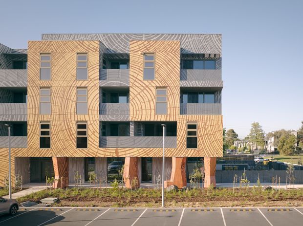 Exterior view of a contemporary multi-storey residential or mixed-use building with distinctive curved, textured timber-toned facade panels, recessed balconies, varied window openings, and ground-level glazing. The building fronts a landscaped verge and marked parking area.