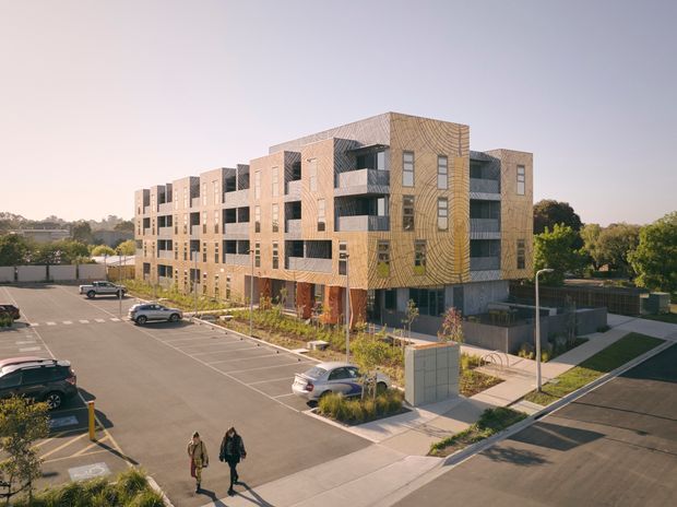 A multi-storey contemporary apartment or mixed-use building viewed from an elevated exterior perspective. The building has a warm timber-toned patterned facade, extensive dark-framed glazing, recessed balconies, and a rooftop or upper-level terrace. It is set within a landscaped urban development with paved walkways, parking areas, low fences, and surrounding trees.
