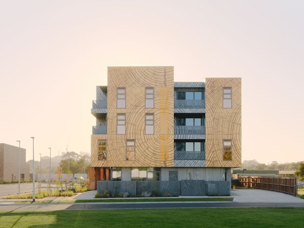 Front exterior view of a contemporary multi-storey residential building with distinctive tan patterned facade panels, dark grey lower-level cladding, recessed balconies, and vertical windows. The building is set within a landscaped site with lawn, pathways, and a driveway or parking area.