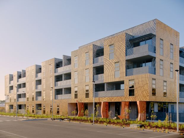 Exterior view of a contemporary multi-storey apartment building with a repetitive stepped facade, projecting balconies, large windows, and warm timber-toned cladding. The building fronts a paved parking area with sparse landscaping and a clear sky.