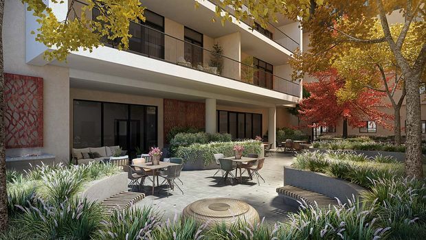 A landscaped communal courtyard or terrace beside a contemporary multi-storey building, furnished with outdoor café tables, chairs, curved lounge seating and a central circular fire pit or table. Dense ornamental planting, mature deciduous trees and apartment balconies frame the paved outdoor space. The architecture features pale rendered walls, dark-framed glazing, projecting balconies and a large decorative red patterned wall panel.