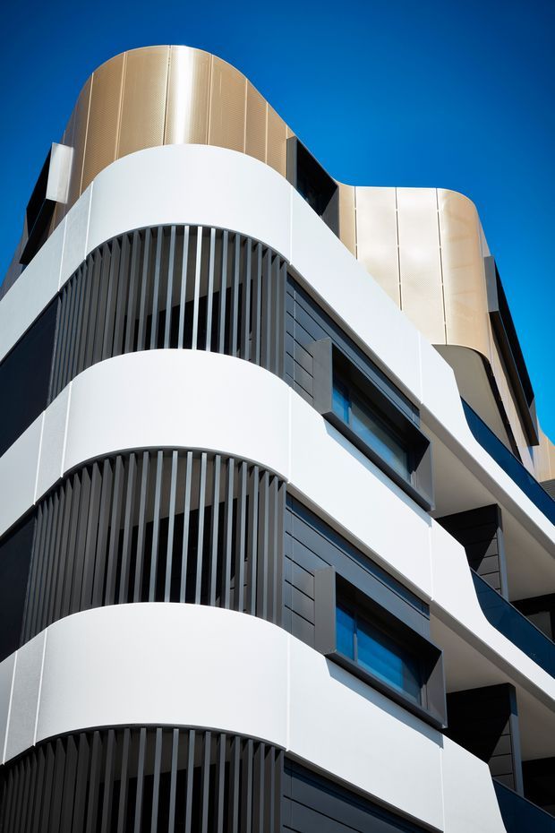 Upward-looking architectural photograph of a contemporary multi-storey building facade. The design features sweeping white curved balcony bands, dark vertical privacy screens, recessed dark window surrounds, blue-tinted glazing, and rounded upper facade volumes clad in a warm metallic or ribbed finish against a clear blue sky.
