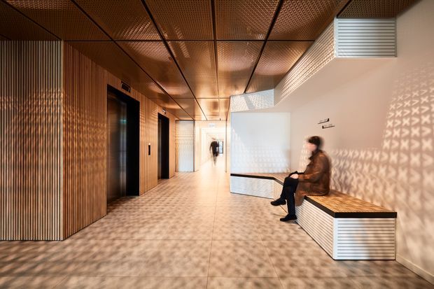 A contemporary interior hallway or lobby with warm timber slat wall cladding, perforated metal ceiling panels, tiled flooring, integrated bench seating, elevator doors, and patterned light projections across the walls and floor. The space has a refined, architectural character with strong linear detailing and warm ambient lighting.