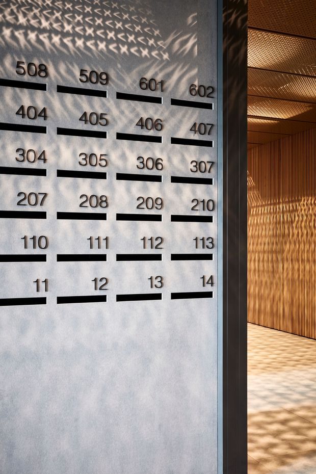 Architectural detail of a modern building entry or lobby. A pale textured wall displays rows of black room numbers with horizontal black nameplate slots, while an adjoining warm timber-lined corridor is visible through a dark metal-framed opening. Patterned sunlight creates strong shadows across the wall, slatted wall, ceiling, and pale tiled floor.