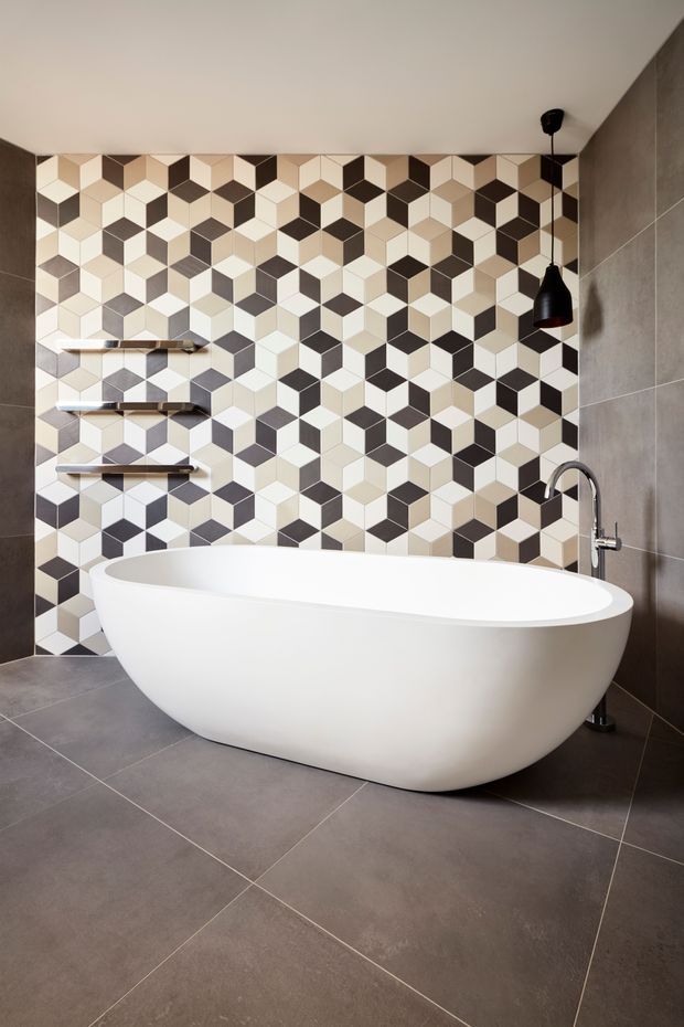 Modern bathroom featuring a large freestanding white oval bathtub, a black and cream geometric patterned tile feature wall, dark grey tiled flooring and side walls, floating shelves, and a black pendant light with a freestanding chrome bath faucet.