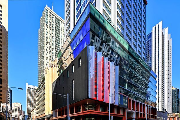 Street-level view of a dense urban commercial building with a dramatic angular glazed facade, dark metal-clad lower volumes, and prominent vertical translucent red and blue feature panels. Multiple tall residential and office towers surround the contemporary structure under a clear blue sky.