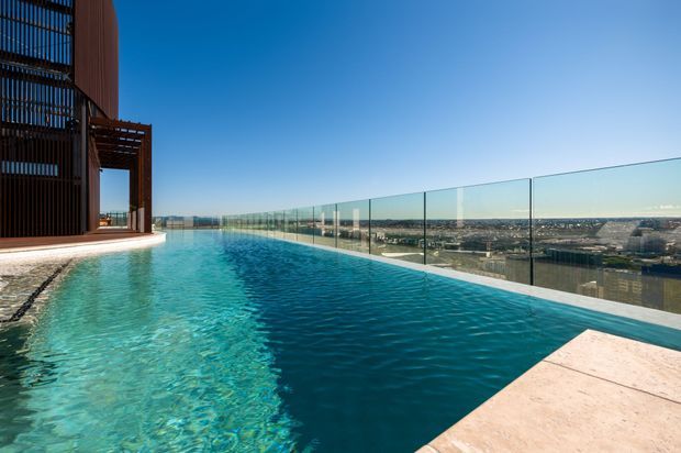 Rooftop infinity pool with clear glass balustrades overlooking a sprawling cityscape. The pool is bordered by pale stone paving and a dark timber-clad architectural structure with vertical screening and a pergola-like projection. Bright blue sky and strong sunlight create a clean, contemporary outdoor setting.