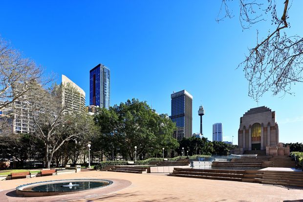 A broad landscaped urban civic plaza with a circular fountain, paved open space, steps, benches, mature trees, and a monumental Art Deco-style stone building. Several modern high-rise office towers rise behind the park, creating a city-centre setting under a clear blue sky.