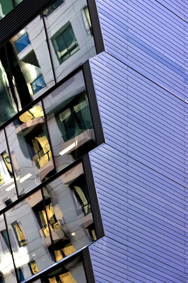 Abstract upward-looking view of a contemporary multi-storey building facade, showing reflective dark-framed glazing and large panels of closely spaced horizontal blue-grey cladding. The angled composition emphasizes the facade geometry, projecting window bays, and strong linear repetition.