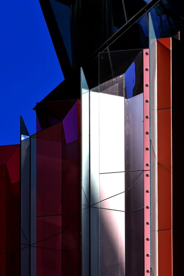 Close-up architectural view of a dramatic geometric facade or sculptural exterior installation. Angular panels and folded planes in deep black, vivid red, magenta, blue, silver, and white create a high-contrast composition, with visible exposed fasteners along a pink-red vertical metal panel. The image is tightly framed against a deep blue sky and emphasizes reflective and opaque facade surfaces rather than an identifiable interior space.