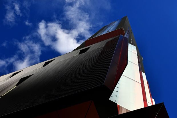 Dramatic low-angle view looking up at a tall contemporary building facade against a vivid blue sky. The structure features sharply angled and projecting forms, extensive dark ribbed metal cladding, silver metallic panels, and bold red and white vertical elements, creating a sculptural high-rise composition.