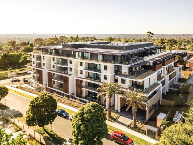 Aerial view of a contemporary multi-storey residential apartment building with a flat roof, layered balconies, extensive glazing, white and dark-toned façades, landscaped grounds, and palm trees. The building sits in an established suburban setting with surrounding low-rise development and tree cover.