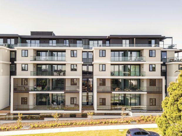 Contemporary multi-storey apartment building facade with a symmetrical arrangement of balconies, recessed glazed living spaces, vertical timber-look screens, dark metal roof elements, and light cream rendered walls. The street-facing landscaping and a partial parked car are visible in the foreground.