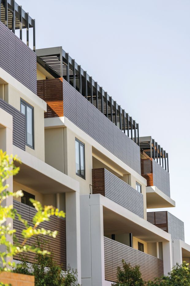 Exterior architectural photograph of a contemporary multi-storey residential apartment or townhouse building. The facade combines pale rendered walls with extensive horizontal dark grey cladding and warm timber-toned privacy screens. Repeated balconies, projecting terraces, tall windows, and rooftop pergola-like shading structures create a strong layered rhythm. Landscaping is visible at the base, with a clear sky behind the building.