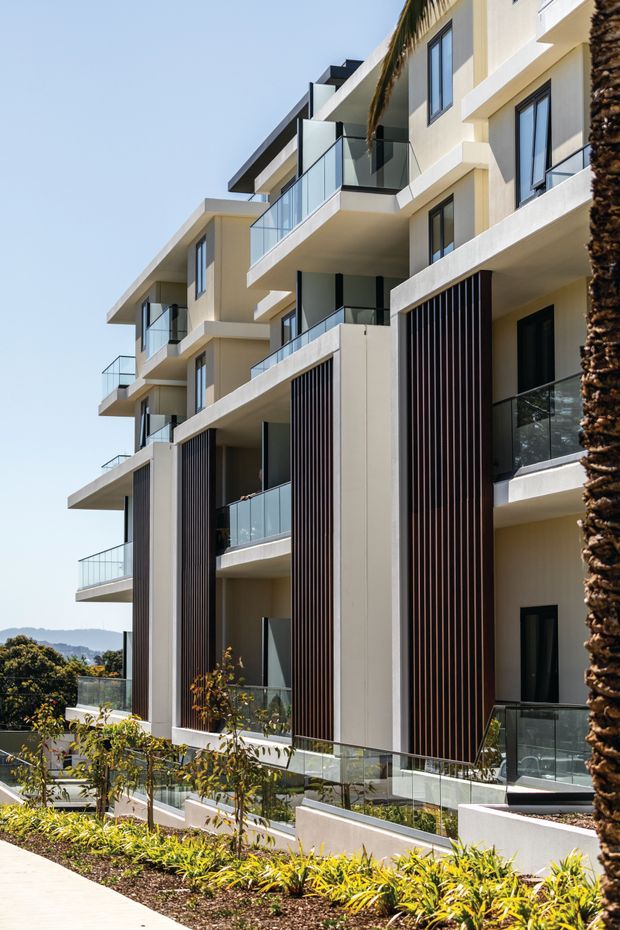 Contemporary multi-storey apartment building exterior with cream rendered walls, projecting balconies, clear glass balustrades, dark-framed windows, and tall vertical dark timber-look screening elements. The landscaped foreground and palm tree reinforce a warm, landscaped urban or suburban setting.