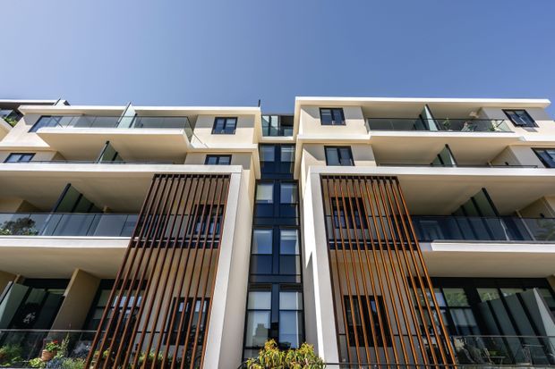 Low-angle exterior view of a contemporary multi-storey apartment building with symmetrical cream-colored balconies, extensive glass balustrades, dark-framed glazing, and prominent vertical timber screens framing the lower levels. The central glazed circulation or atrium zone emphasizes the building's modern residential facade.