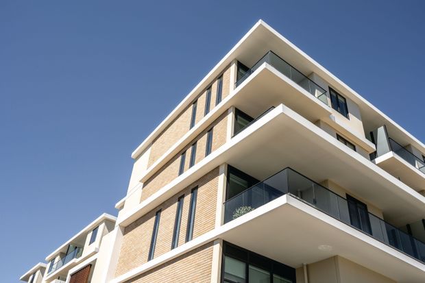 Low-angle exterior view of a contemporary multi-storey apartment building with stepped projecting balconies, dark glazed balustrades, cream and light tan brick facade panels, and large dark-framed windows against a clear blue sky.