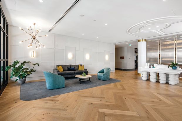 A bright, refined contemporary lobby or reception lounge with a large open seating area. The room features a warm herringbone timber floor, pale geometric wall panelling, recessed ceiling lights, a sculptural brass-toned starburst pendant, a dark sofa with yellow cushions, teal lounge chairs, and a low round coffee table on a large grey rug. At right is a white reception or service counter with cylindrical supports, backed by a decorative circular ceiling feature and screened glazing.