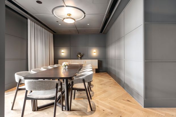 A refined contemporary dining room featuring a long dark timber dining table surrounded by upholstered dining chairs. The room has grey wall panelling, integrated cabinetry, full-height sheer curtains, warm wall sconces, a sculptural circular ceiling light, and light timber herringbone flooring.