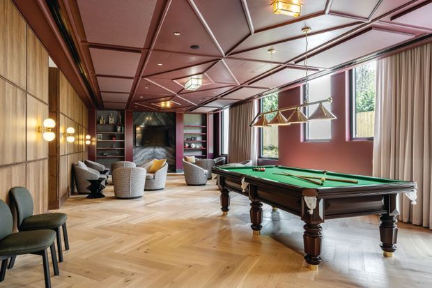 A refined billiards room with a full-size green-felt pool table, brass billiard lighting, and upholstered lounge chairs. The room features warm herringbone timber flooring, timber wall panelling, rich burgundy walls, geometric coffered ceiling detailing, and floor-to-ceiling curtains with generous windows. Built-in shelving and a dark fireplace or feature wall add depth to the sophisticated interior.