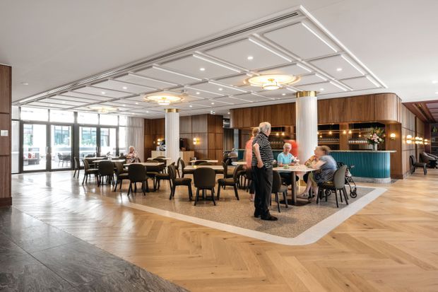 A spacious, upscale hotel or hospitality lounge with a restaurant and bar setting. The room features a decorative coffered ceiling with integrated downlights and circular pendant fixtures, fluted white columns, dark timber wall panelling and cabinetry, a curved teal bar, patterned carpet seating zones, and pale timber herringbone flooring. Multiple dining tables and lounge chairs are arranged around the open-plan space, with large glazed doors and windows at the rear.