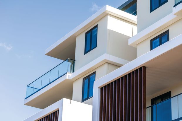 Close-up exterior view of a contemporary multi-level residential building with crisp white rendered walls, projecting flat roof slabs and balconies, dark-framed blue-tinted glazing, glass balustrades, and vertical timber screening. The composition highlights modern facade articulation and layered volumes against a clear sky.