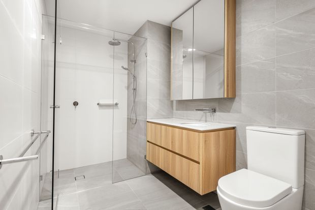 Bright contemporary bathroom with a frameless glass walk-in shower, wall-mounted timber vanity with integrated white basin, mirrored cabinet, wall-hung towel rail, and white toilet. Large pale grey stone-look tiles cover the walls and floor, creating a clean, refined finish.
