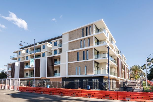 Exterior street view of a contemporary multi-storey apartment building under construction or near completion. The building features light brick and white rendered facade sections, projecting balconies with glass balustrades, tall narrow windows, dark vertical screening elements, and a flat roof. Red construction barriers and temporary fencing line the foreground.