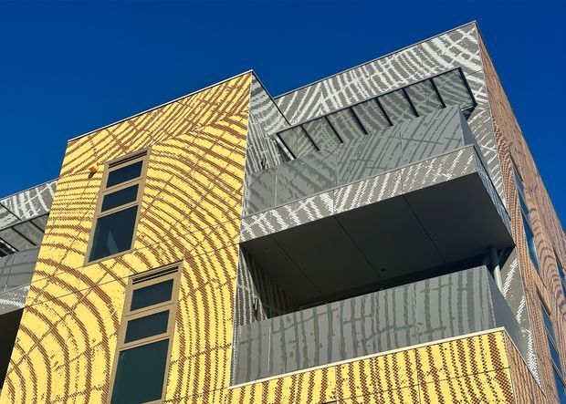 Low-angle exterior view of a multi-storey contemporary building with bold patterned facade panels in yellow, grey, white, and brick tones. The facade features projecting balconies, dark metal or aluminium-framed windows, and deep balcony soffits against a clear blue sky.