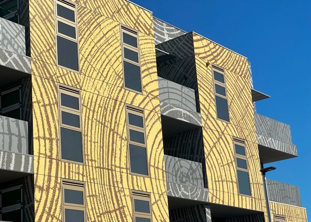Exterior view of a contemporary multi-storey building with a striking yellow patterned facade, narrow vertically aligned windows, projecting balconies, and contrasting grey patterned panels. The geometric massing and balcony forms create a bold urban architectural composition against a clear blue sky.
