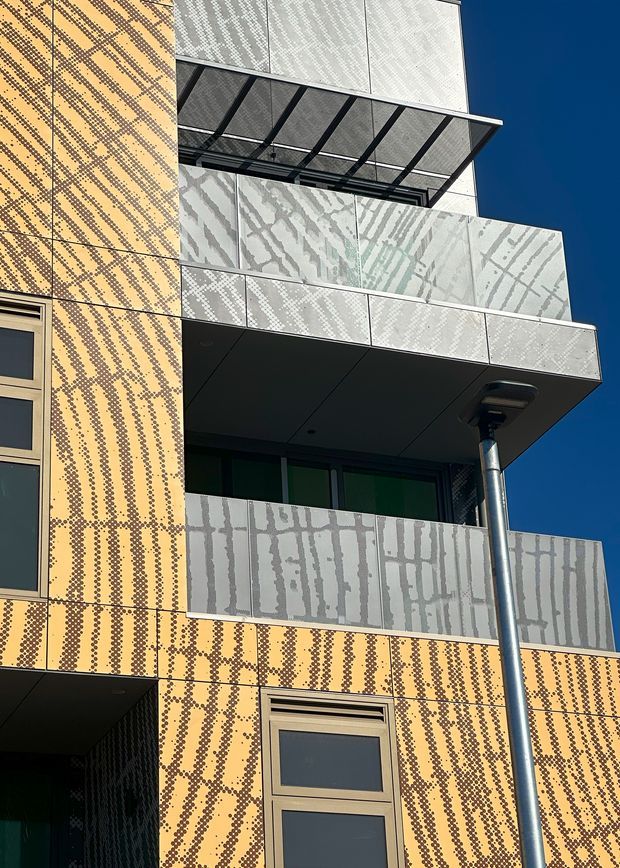 Close-up exterior view of a contemporary multi-storey building facade featuring patterned yellow-orange cladding, pale perforated or patterned metal balcony panels, dark metal balcony canopies, glazed doors and windows, and a projecting balcony. A streetlight is visible in the foreground against a clear blue sky.