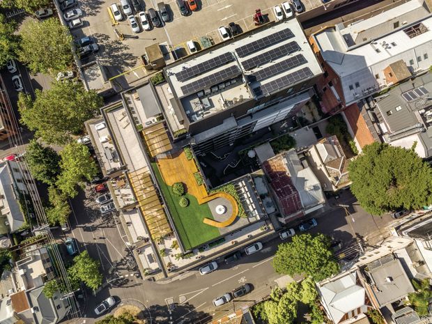 Aerial view of a dense urban mixed-use or commercial building complex surrounded by streets and parked cars. The central flat-roofed building has extensive rooftop solar panels, a rooftop green terrace with timber decking, curved lawn areas, and a circular outdoor feature. Adjacent low-rise buildings, mature street trees, and a parking area are also visible.