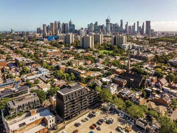 Aerial panoramic view of a dense urban neighbourhood with a modern high-rise skyline in the background, surrounded by low-rise residential buildings, mature trees, streets, and a surface parking area. The image primarily documents the cityscape and urban setting rather than a single building.