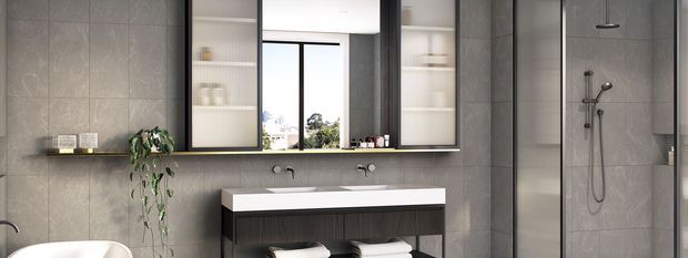 Contemporary grey-tiled bathroom with a long double vanity, white integrated basin, dark timber cabinetry, wall-mounted taps, large mirrored storage, open shelving, and a glazed walk-in shower. A freestanding or partially visible white bathtub appears at the left edge. The room has restrained monochrome finishes, black-framed glazing and fittings, and subtle greenery.