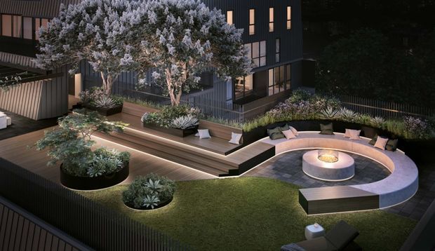A nighttime architectural rendering of a landscaped rooftop or elevated outdoor terrace beside a dark contemporary building. The space features timber decking, planted garden beds and trees, a sunken circular lounge with curved built-in seating around a central fire feature, and warm integrated landscape lighting.