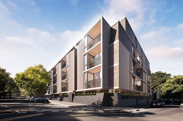 Exterior architectural photograph of a contemporary multi-storey apartment building on an urban, tree-lined corner site. The building has a strong angular pitched/gabled roof form, projecting balconies, large glazed openings, vertical dark metal cladding, pale brick masonry, and a dark masonry or concrete podium. Cars and street landscaping are visible around the site.