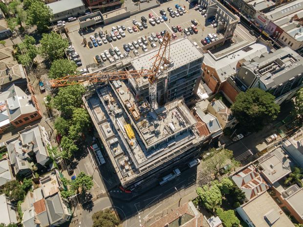 Aerial view of a dense urban construction site with a multi-storey building under construction, tower crane, exposed concrete structure, surrounding streets, low-rise buildings, mature trees, and a nearby surface car park.