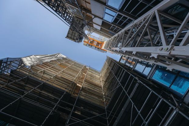 Upward-looking view between high-rise buildings under construction, showing extensive steel scaffolding, temporary structural framing, construction hoists, cranes, exposed facade systems and blue sky. The image documents an active urban construction site rather than a completed interior or exterior facade.