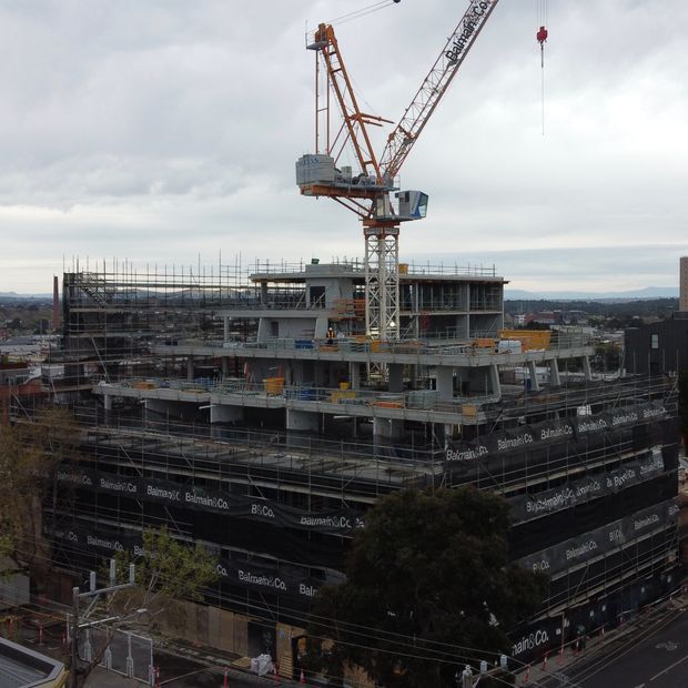 A multi-storey concrete building under construction in an urban setting, surrounded by extensive scaffolding and safety mesh. A large tower crane dominates the scene, with exposed structural floors, temporary construction platforms, and distant low-rise development visible under an overcast sky.