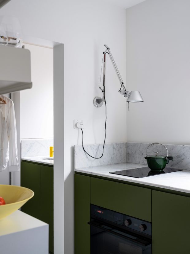 Compact contemporary kitchen with dark green lower cabinetry, white marble-look benchtops and splashback, integrated black oven, induction cooktop, and a chrome articulated wall-mounted lamp. A partial adjoining laundry or utility area is visible through the opening on the left.