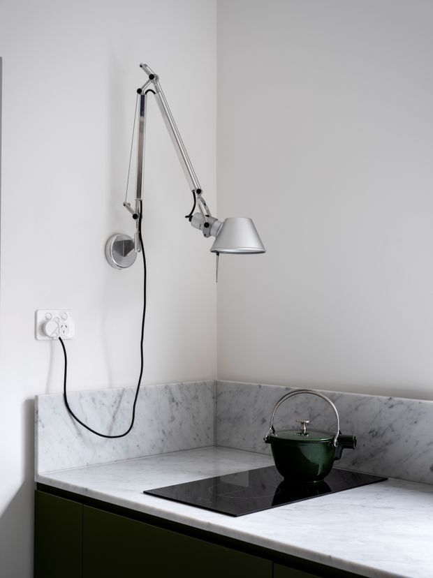 A compact, minimalist kitchen corner with a white veined marble benchtop and matching low splashback. Dark green lower cabinetry supports a black induction cooktop, with a deep green kettle placed on top. A silver articulated wall-mounted task lamp and exposed power cord add a utilitarian detail against the plain white walls.