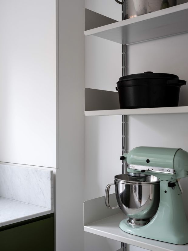 Close-up of a neatly organised kitchen storage area with open white shelves holding a black cooking pot and a mint-green stand mixer with a stainless-steel bowl. White cabinetry and a pale marble backsplash are visible, with a dark green lower cabinet edge at the bottom left. The image has a clean, contemporary minimalist appearance with a muted, cool-toned palette.