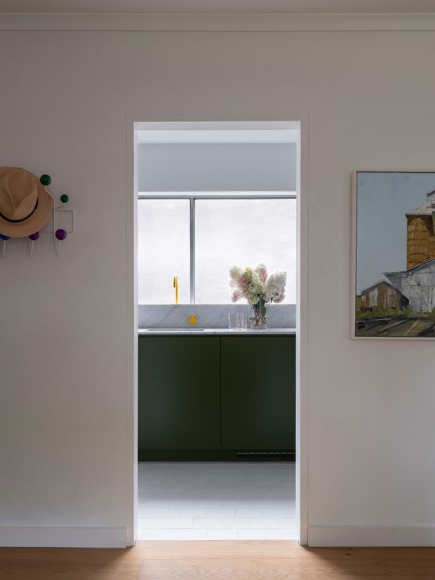View from a softly lit hallway or living area through a doorway into a compact kitchen. The kitchen features dark green flat-panel cabinetry, a pale stone or composite benchtop, a large window with a grey roller blind, and light tiled flooring. The surrounding room has warm neutral painted walls, timber flooring, simple white trim, a wall-mounted hat rack, and framed artwork.