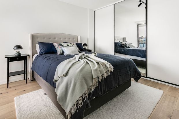 A bright, contemporary bedroom with a large upholstered bed dressed in dark navy bedding, layered pillows, and a light fringed throw. Full-height mirrored wardrobe doors line one wall, reflecting the bed and window. Pale timber flooring, white walls, a neutral rug, and a small black bedside table with lamp create a clean, restrained interior.