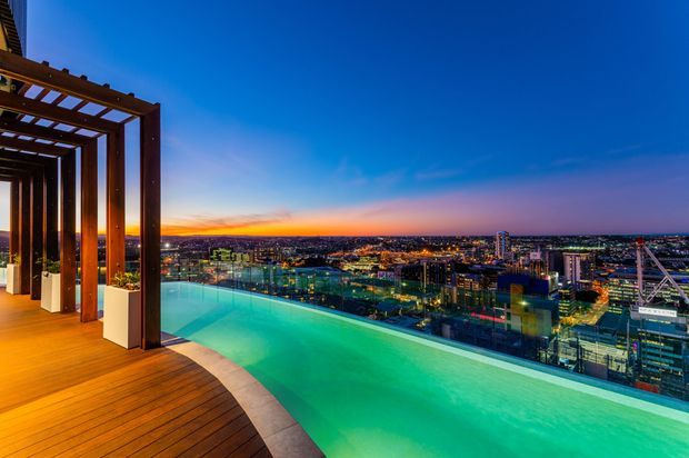 A rooftop terrace at dusk with a glowing infinity pool overlooking a dense illuminated city skyline. The pool is bordered by a timber deck and a dark timber pergola with integrated planter boxes, creating a luxurious elevated outdoor entertaining space.