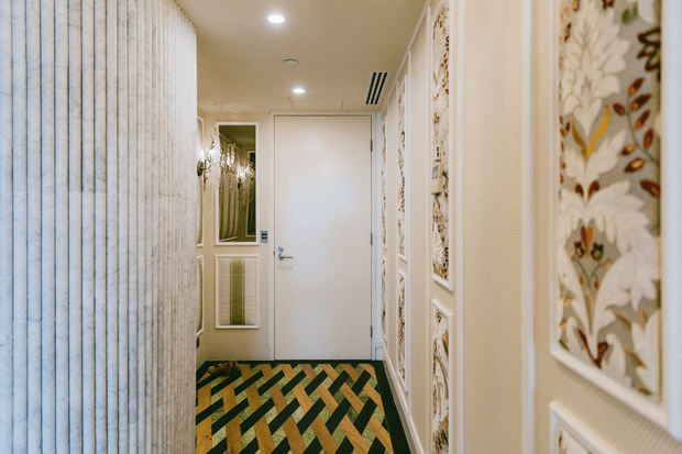 Narrow residential hallway or entry corridor with a closed white door, patterned geometric carpet, textured vertical wall covering on the left, and ornate floral decorative wall panels on the right. Recessed ceiling lights and a wall mirror contribute to a refined, decorative interior.