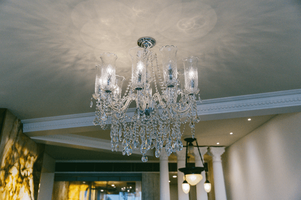 Interior view dominated by an ornate crystal chandelier hanging from a detailed ceiling with decorative crown moulding. Recessed ceiling lights, classical columns, pendant lights, and a warm-toned textured feature wall are visible in the background, suggesting a refined, traditionally styled foyer or living space.