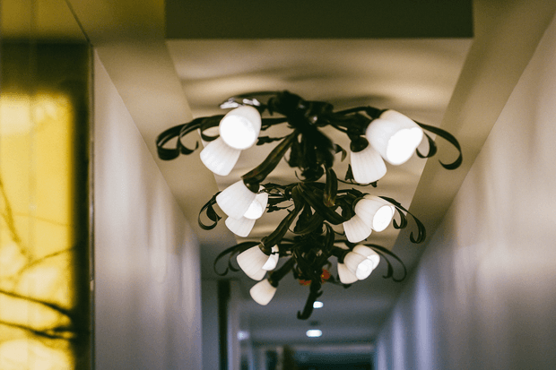 A narrow interior hallway viewed beneath an ornate, multi-arm chandelier with dark scrolling metalwork and numerous white glass shades. The corridor has pale walls and a low ceiling, with warm ambient lighting and a softly blurred background.